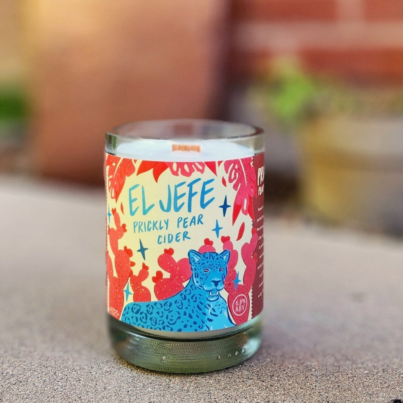 A recycled glass candle crafted from a cider bottle, featuring a bold and colorful label for "El Jefe Prickly Pear Cider." The label displays vibrant red cactus motifs and a striking blue mountain lion illustration with playful text. The candle rests on a concrete surface outdoors, with a blurred background of potted plants and a red brick wall, evoking a lively and Southwestern-inspired atmosphere. The smooth glass rim and creamy white wax enhance its polished and artisanal look