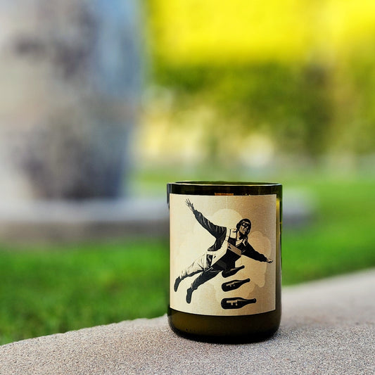 A recycled glass candle made from a green wine bottle, featuring a minimalist label with a striking black-and-white illustration of a pilot in freefall alongside bottle-shaped silhouettes. The candle sits outdoors on a smooth, light stone surface, with a blurred background of vibrant green grass and abstract shapes, evoking a modern, adventurous aesthetic.