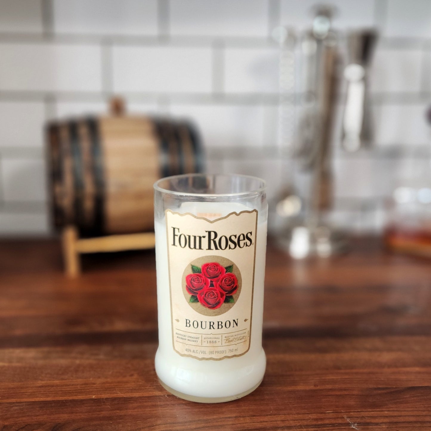  A recycled Four Roses Bourbon bottle transformed into a candle, featuring creamy white wax and the iconic label with red roses. The candle is displayed on a wooden countertop with a mini bourbon barrel and cocktail tools blurred in the background, creating a cozy, rustic ambiance.