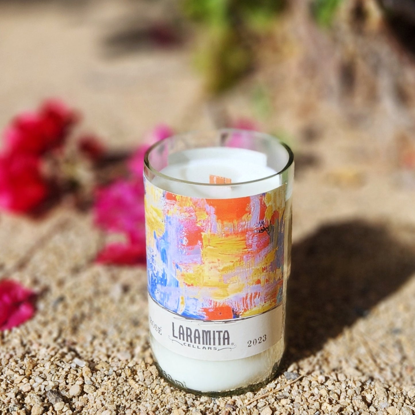 A handcrafted candle in a recycled glass vessel made from a Laramita Cellars wine bottle, featuring an abstract, colorful label with vibrant strokes of blue, orange, yellow, and pink. The candle, with a creamy white wax and wooden wick, is set on sandy ground, surrounded by blurred pink bougainvillea flowers and greenery, creating a bright and artistic desert-inspired ambiance.