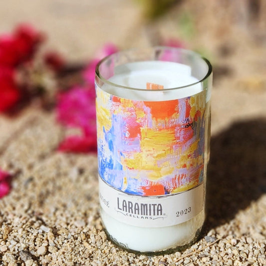 A handcrafted candle in a recycled glass vessel made from a Laramita Cellars wine bottle, featuring an abstract, colorful label with vibrant strokes of blue, orange, yellow, and pink. The candle, with a creamy white wax and wooden wick, is set on sandy ground, surrounded by blurred pink bougainvillea flowers and greenery, creating a bright and artistic desert-inspired ambiance.