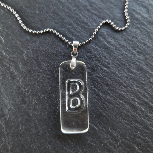 Handmade Letter B initial necklace featuring original raised lettering in clear recycled whiskey bottle glass, with a stainless steel bail and gunmetal ball chain on textured charcoal slate.