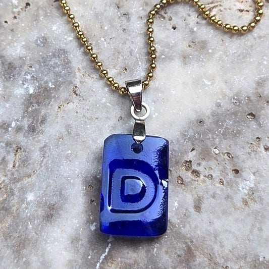 Cobalt-blue recycled kombucha bottle glass Letter D necklace with original raised lettering, a stainless steel bail, and gold-tone ball chain on textured travertine.