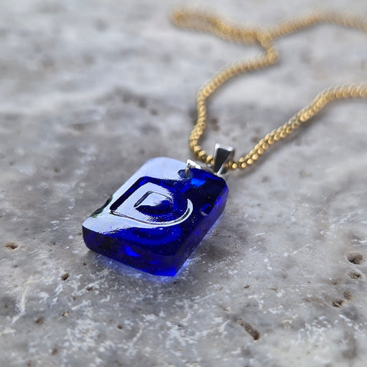 Low-angle close-up of a cobalt-blue Letter D pendant handcrafted from reclaimed bottle glass, showing the molded raised letter, polished edges, and glass thickness.