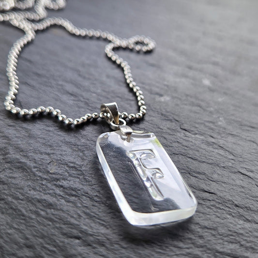 Low-angle close-up of a reclaimed clear bottle glass Letter F pendant, highlighting the original raised lettering, thick polished glass edges, and silver-tone chain against textured slate.