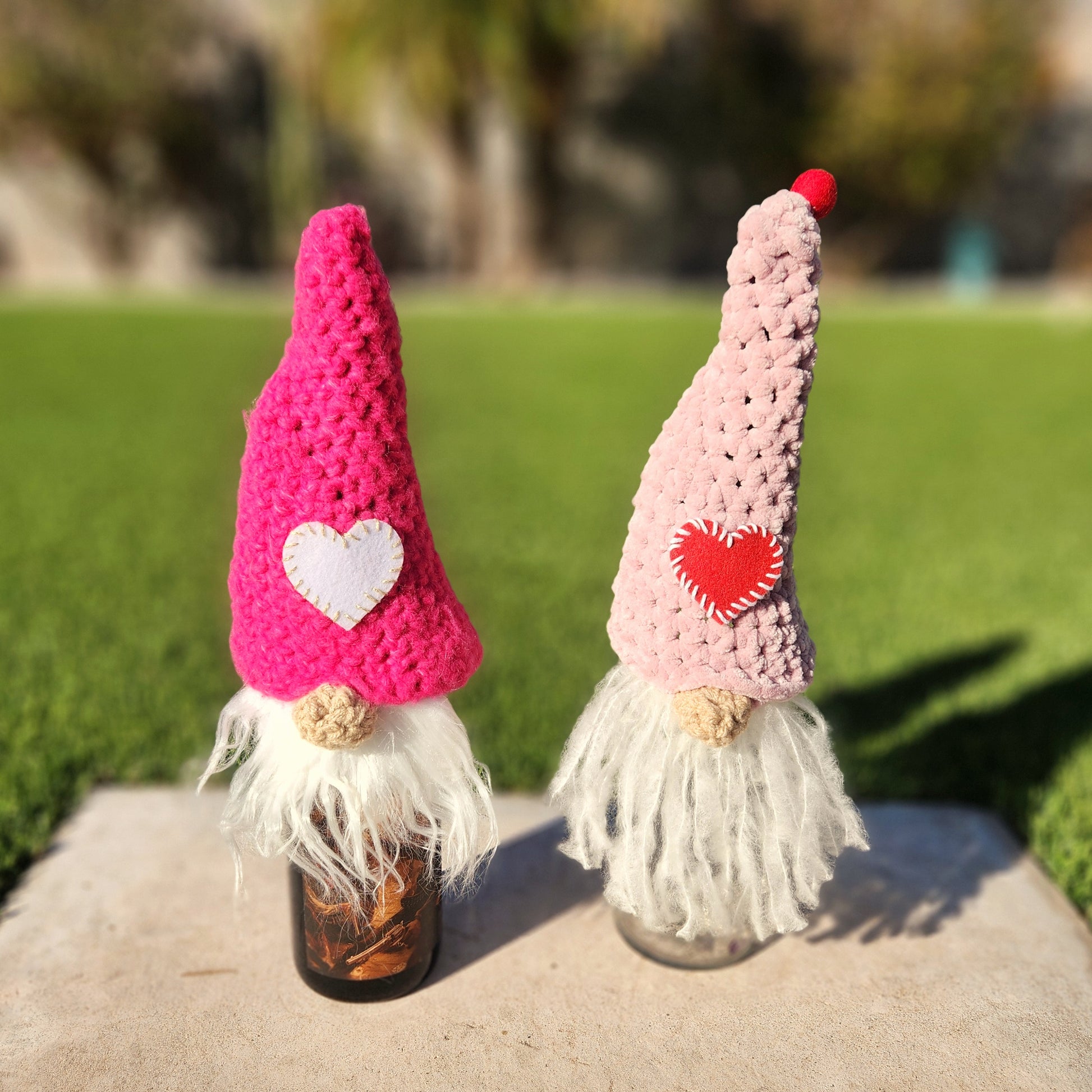 Two handcrafted crochet gnomes, adorned with long white beards. One gnome features a bright pink hat with a white felt heart, and the other has a pale pink hat with a red stitched heart and a pom-pom on top. They are displayed on a concrete surface with a vibrant green lawn and blurred background, creating a whimsical and eco-friendly design aesthetic.