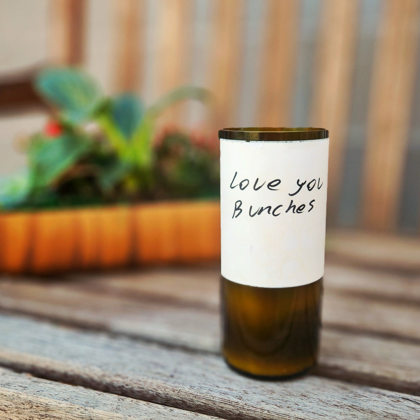 A recycled wine bottle candle with a clean-cut, smooth glass rim, featuring a handwritten-style label that reads "Love you bunches." The candle is placed on a wooden bench with a blurred background of greenery and a planter, creating a cozy and heartfelt ambiance.