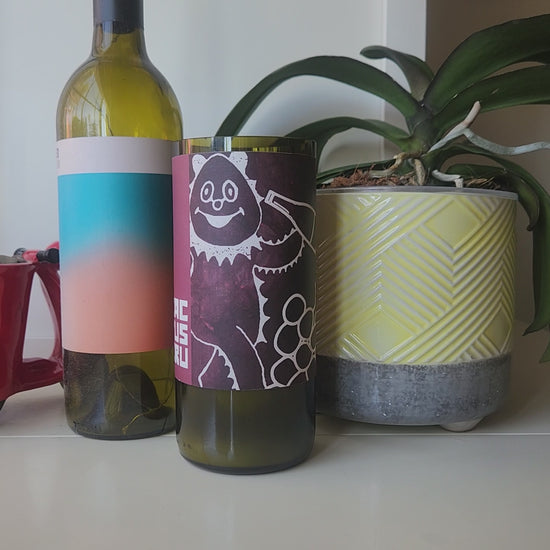 Video of playful burgundy wine bottle candle being spun to show all sides, including the smooth wax top and wooden wick.  Full wine bottle and plant are in the background.