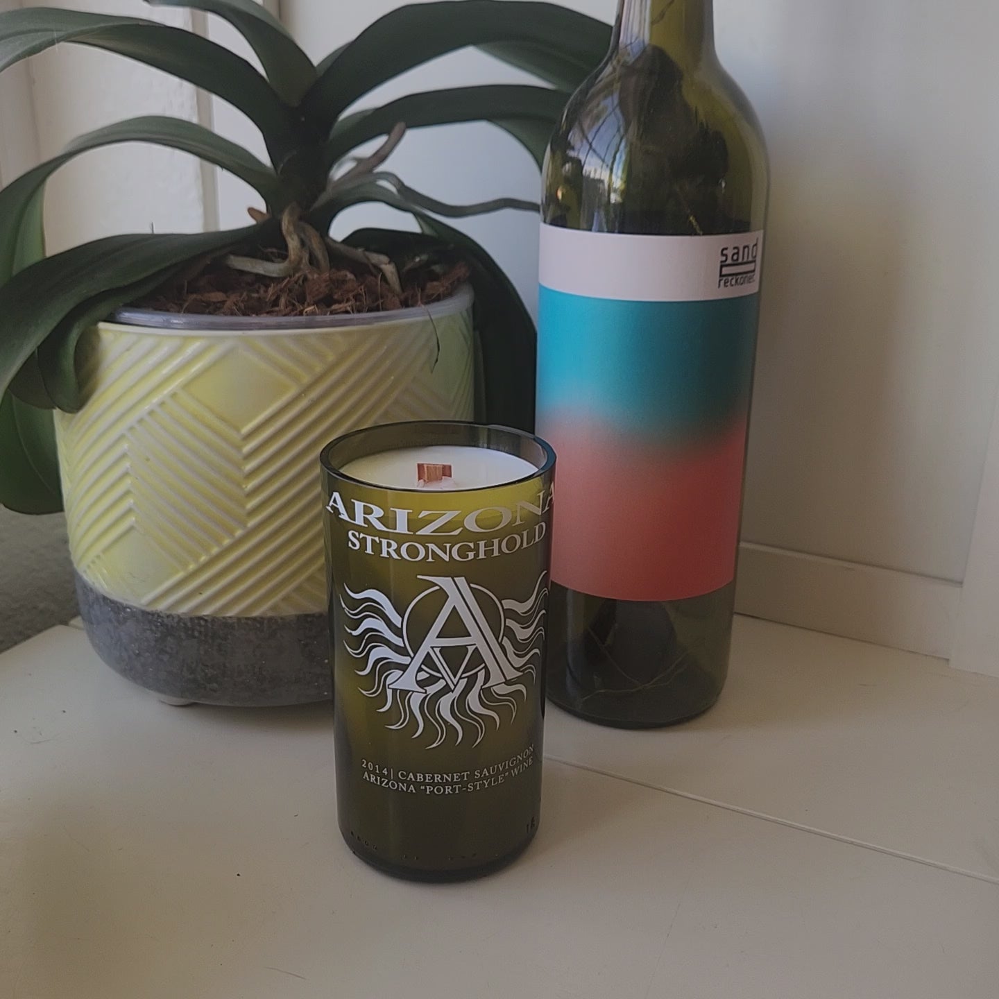 Video of Arizona Stronghold wine bottle candle being spun around, showcasing its smooth shiny rim, eco friendly wax, and wooden wick.  Plant and wine bottle in the background.