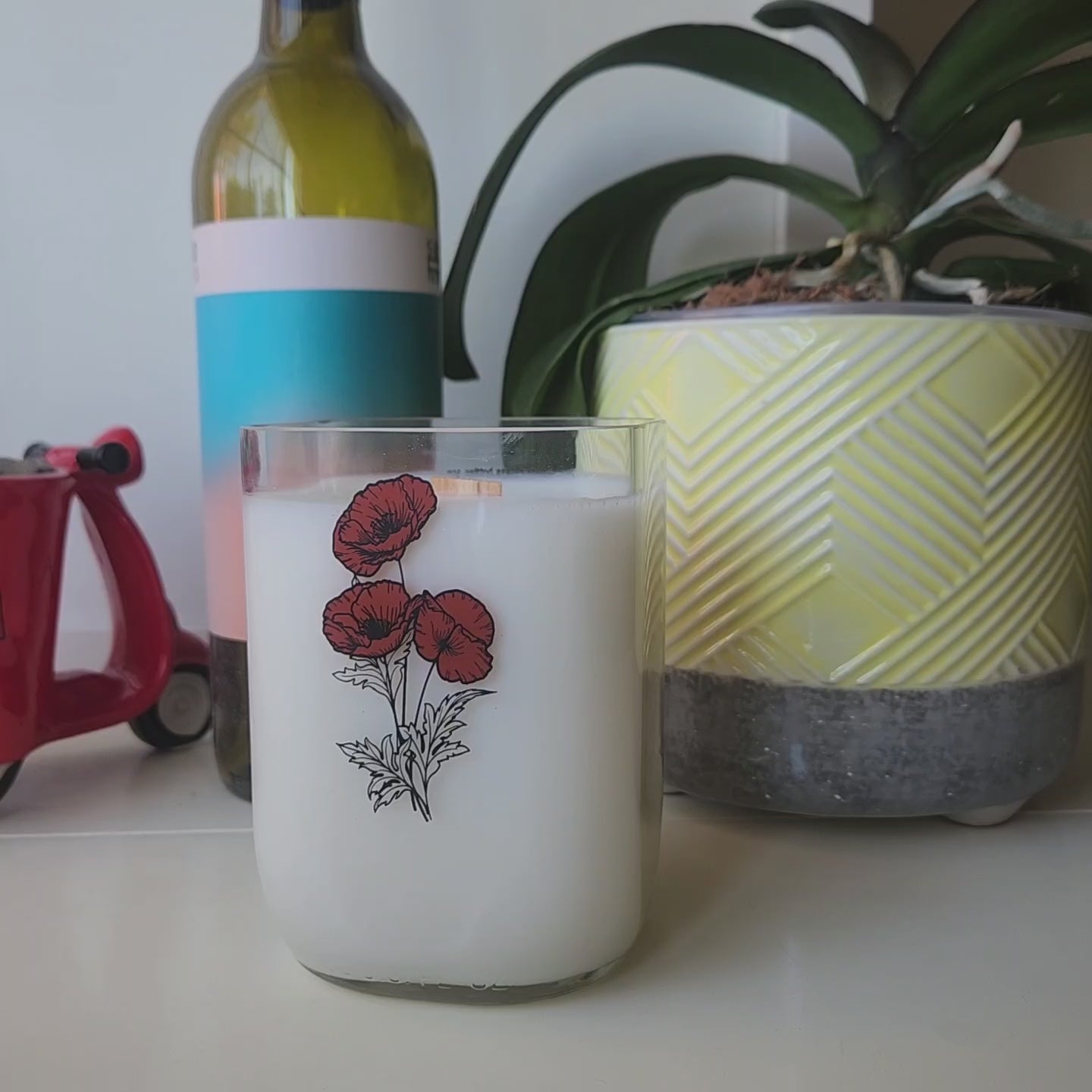 Video of milk bottle candle being spun around to see all sides, including the smooth wax top and wooden wick.  Wine Bottle and plant are in the background.