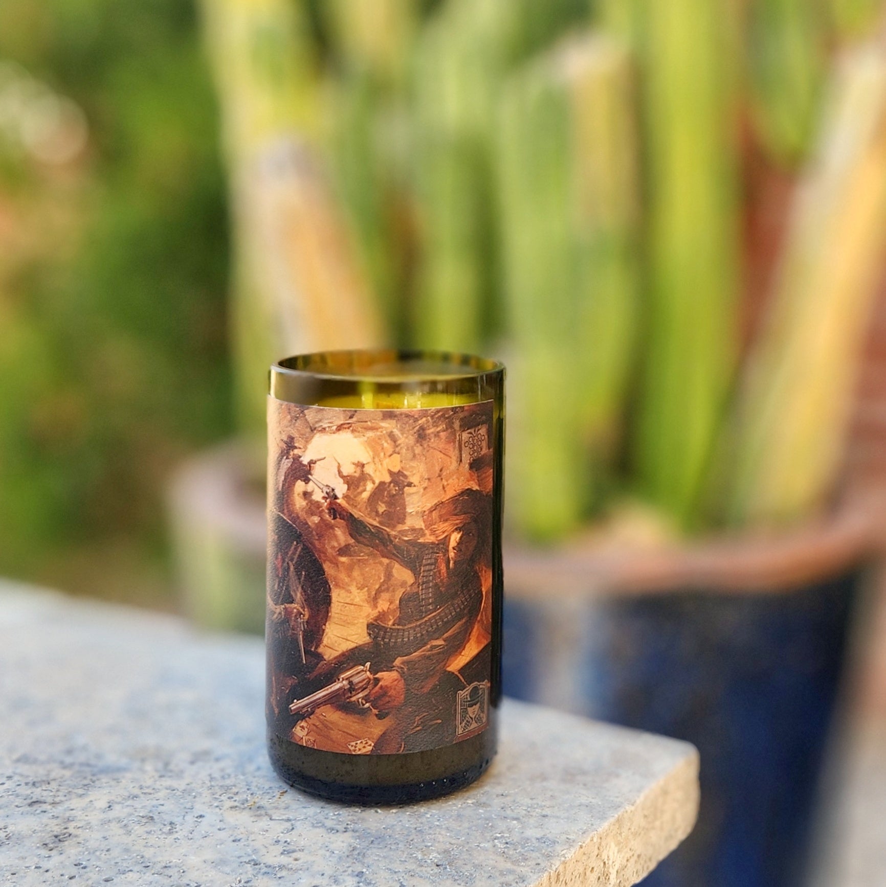 A recycled wine bottle candle with a dramatic illustrated label depicting a vintage Western-style scene. The candle is placed on a textured stone surface with a blurred background of tall potted cacti and a brick wall, creating a rustic desert ambiance.