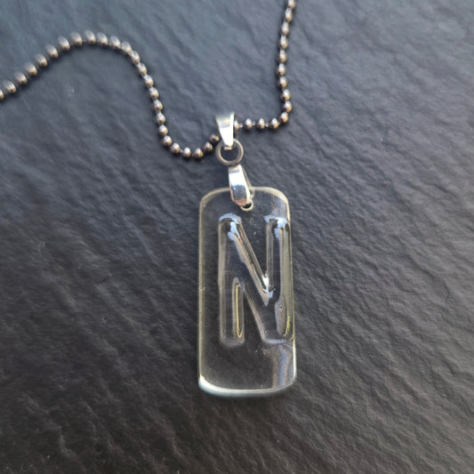 Close-up of a handmade recycled bottle glass initial necklace featuring a clear pendant with a raised letter N, stainless steel bail, and gunmetal ball chain. Photographed on dark gray slate.