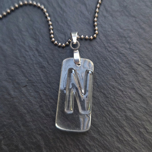 Close-up of a handmade recycled bottle glass initial necklace featuring a clear pendant with a raised letter N, stainless steel bail, and gunmetal ball chain. Photographed on dark gray slate.