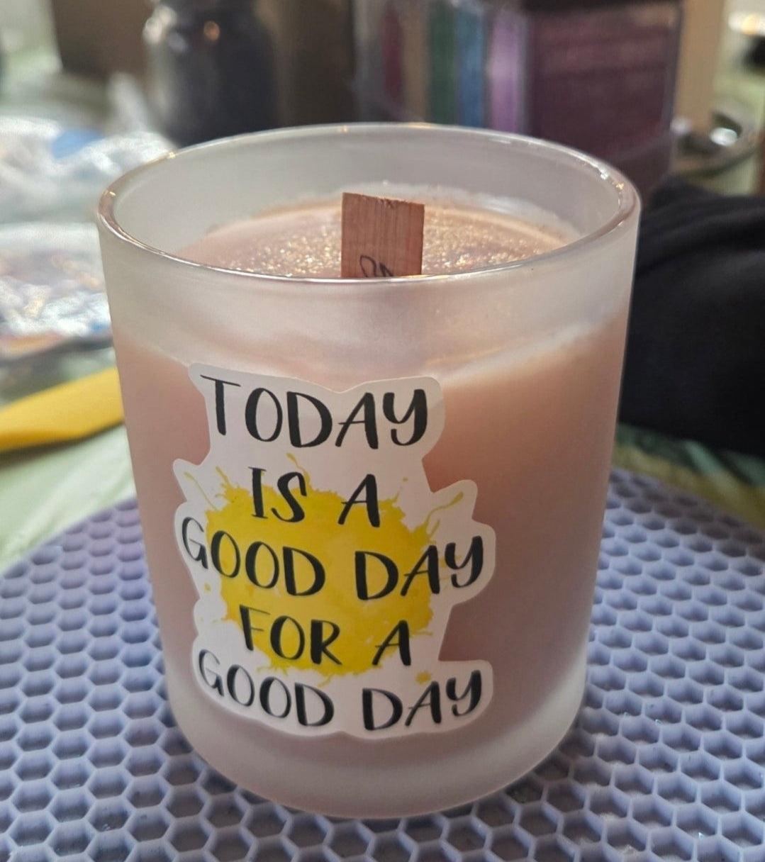 Candle in a glass holder with a label reading 'Today is a good day for a good day' on a textured surface.