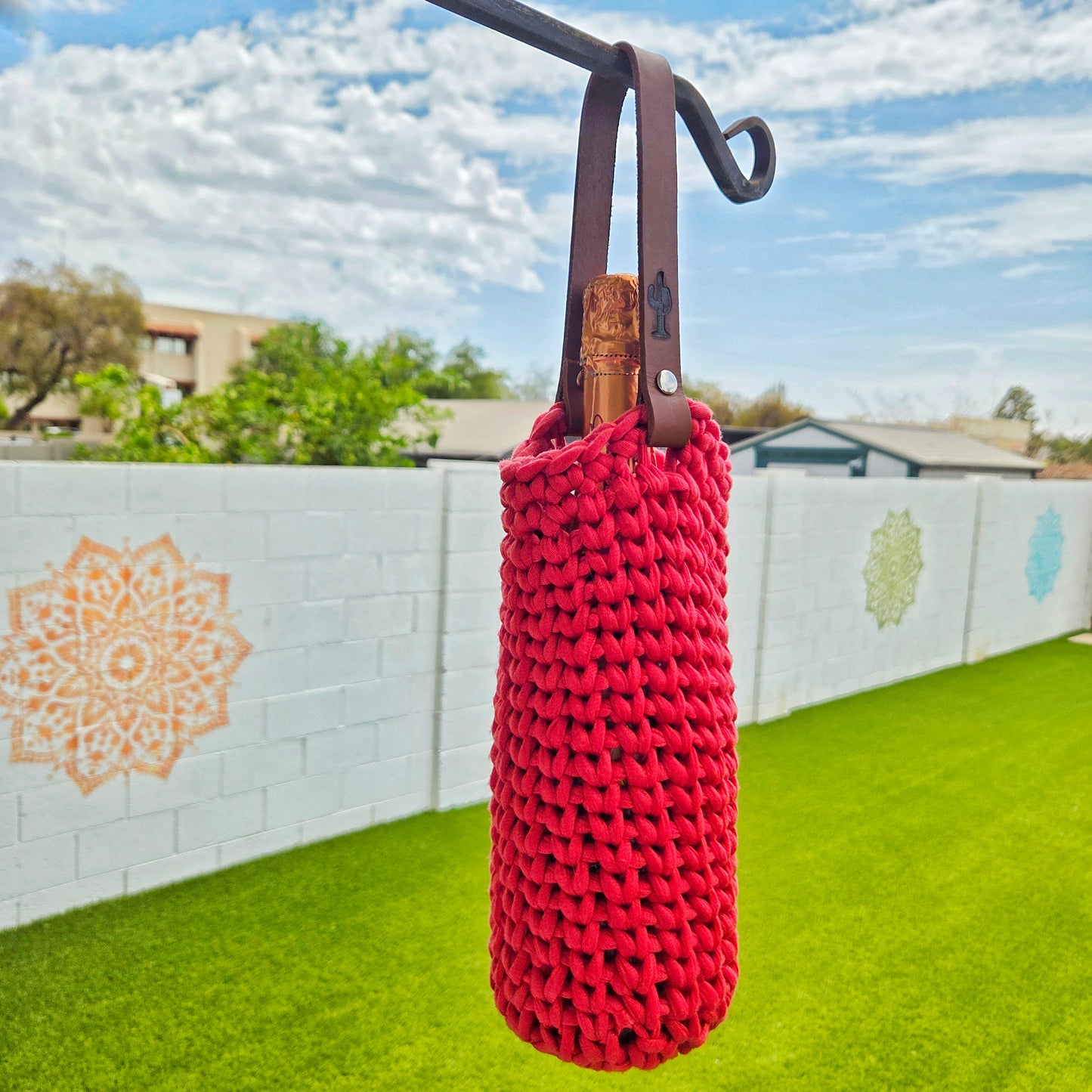 A bright red hand-knit wine bottle sleeve with a dark brown leather handle, holding a wine bottle, hanging from a black metal hook in an outdoor backyard setting with a white brick wall, green grass, and blue sky.