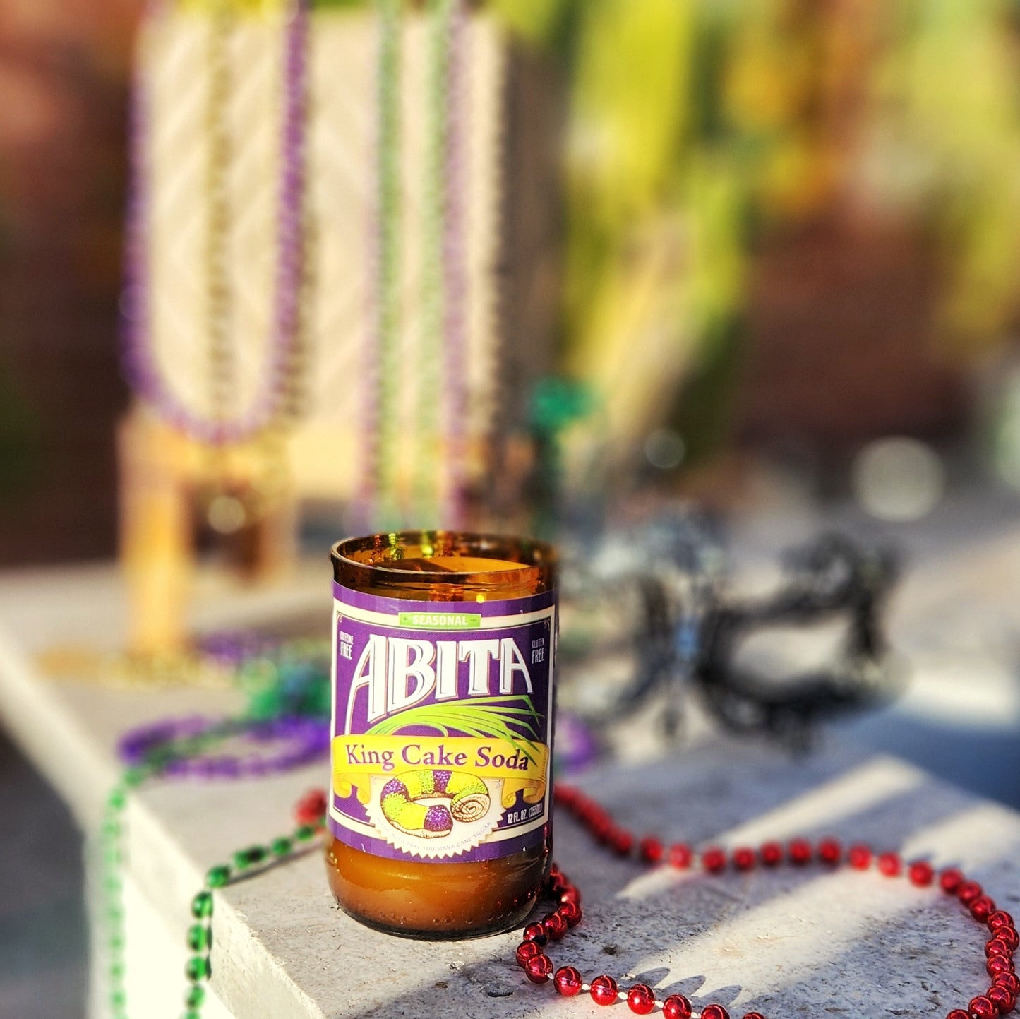 A hand-poured coconut soy wax candle made from a recycled Abita King Cake Soda bottle. The festive label showcases bright Mardi Gras colors with an illustration of a King Cake, celebrating the spirit of New Orleans. This eco-friendly wooden wick candle has a polished glass rim, highlighting its artisanal craftsmanship. Placed on a sunlit stone surface surrounded by colorful Mardi Gras beads, this upcycled soda bottle candle is perfect for fans of New Orleans culture and sustainable gifts.