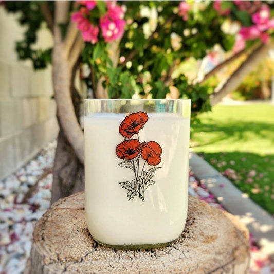 Delight in the charm of this hand-poured candle crafted in a recycled milk bottle, featuring a delicate poppy design. Set on a rustic tree stump with vibrant blooms in the background, this piece brings eco-friendly elegance to any space.