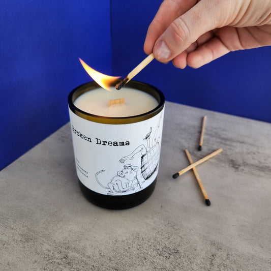 White candle with wooden wick made in a cut off wine bottle.  Label shows a drawing of a monkey and a barrel with legs coming out of it.  Label says "Broken Dreams".  Candle is being lit.