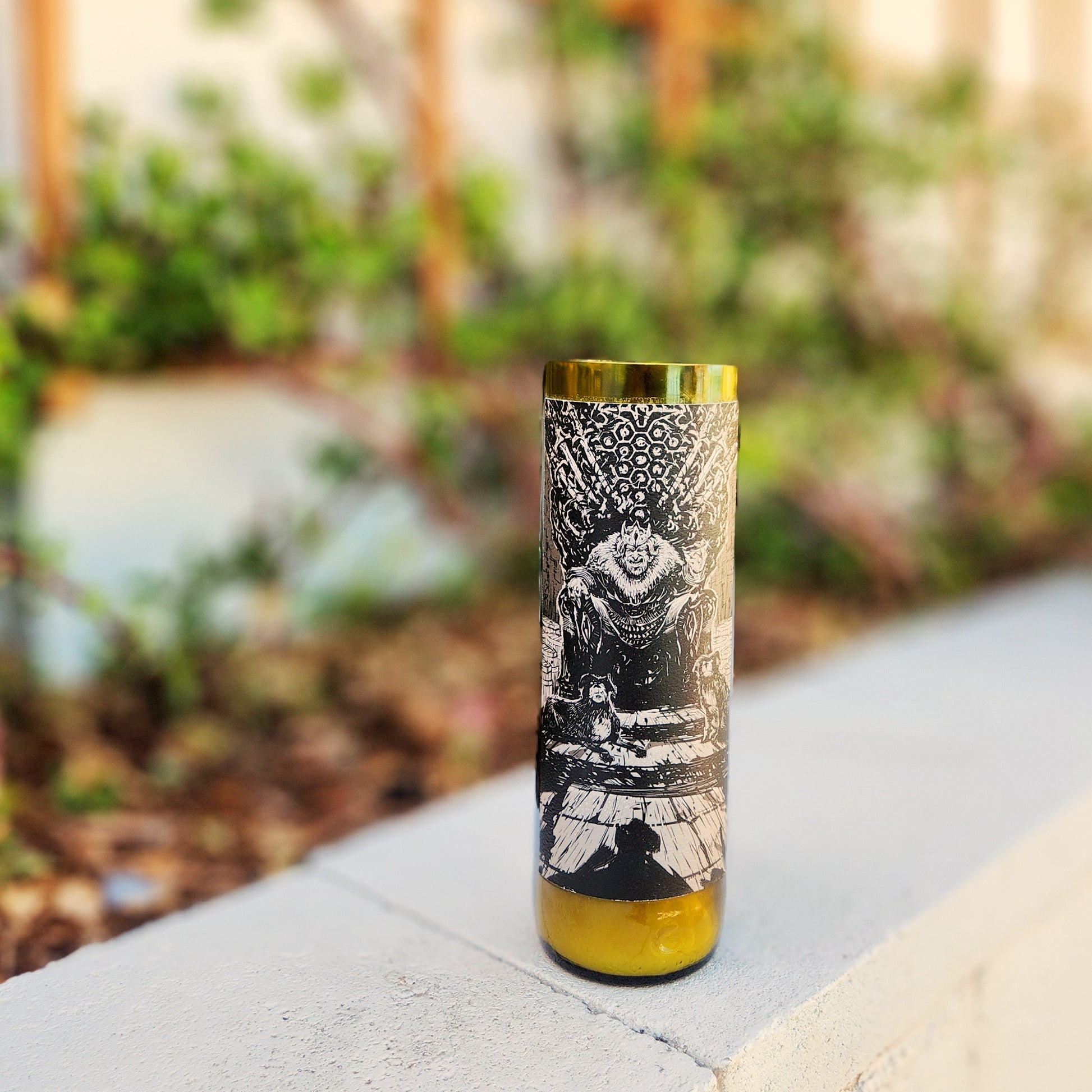 A recycled glass candle crafted from a green wine bottle, featuring an intricate black-and-white label with an imposing illustration of a king-like figure seated on a throne adorned with ornate details. The label's dark and moody design adds a regal and mysterious vibe. The candle is placed outdoors on a light concrete surface, with blurred greenery in the background, enhancing its bold and striking appearance.