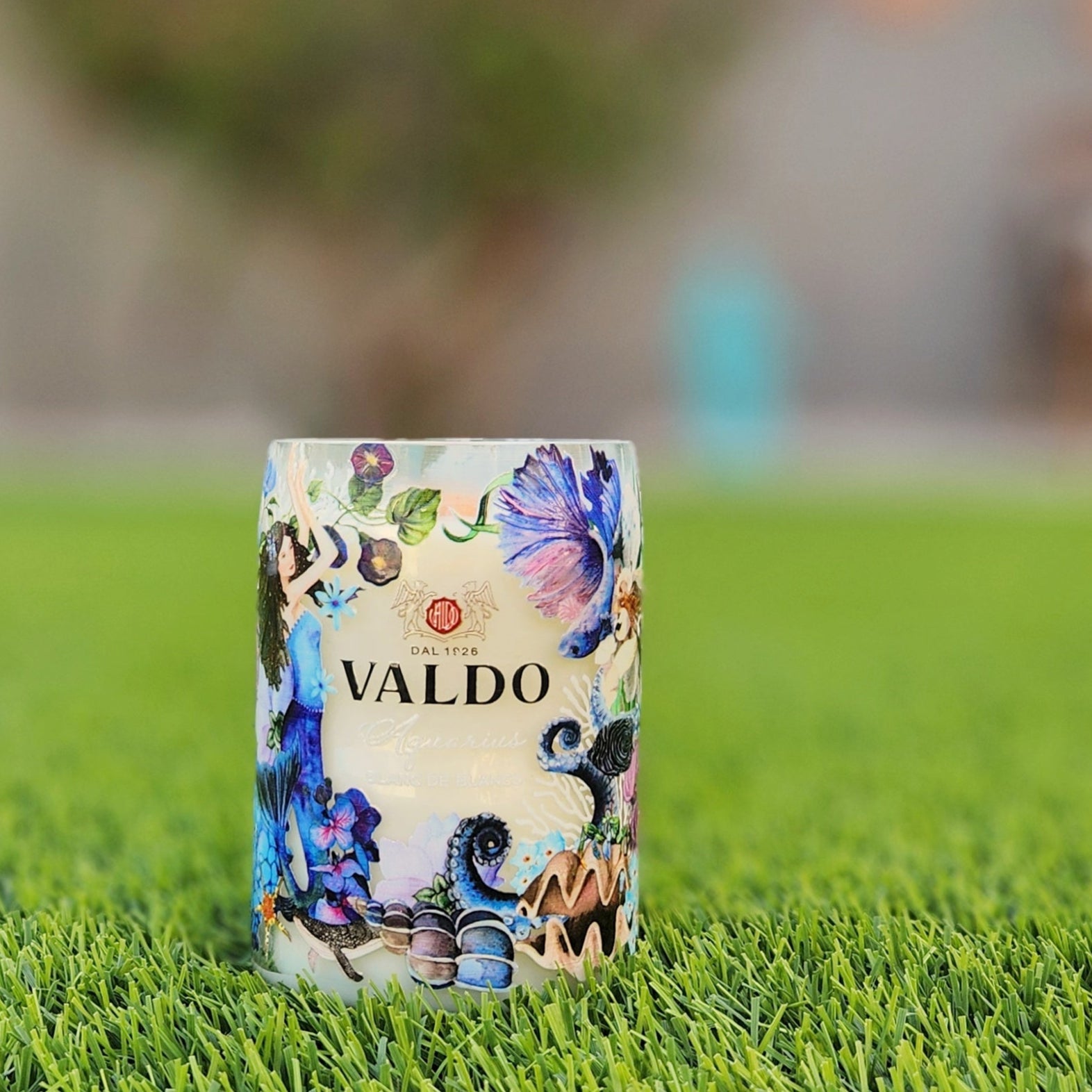 A handcrafted candle made from a recycled Valdo sparkling wine bottle, featuring an intricate and artistic label with floral and marine-inspired illustrations in shades of blue, purple, and gold. The candle sits on a vibrant green lawn, with a softly blurred background of warm glowing lights, a bench, and greenery. The polished glass rim and creamy wax highlight its eco-friendly craftsmanship and luxurious aesthetic