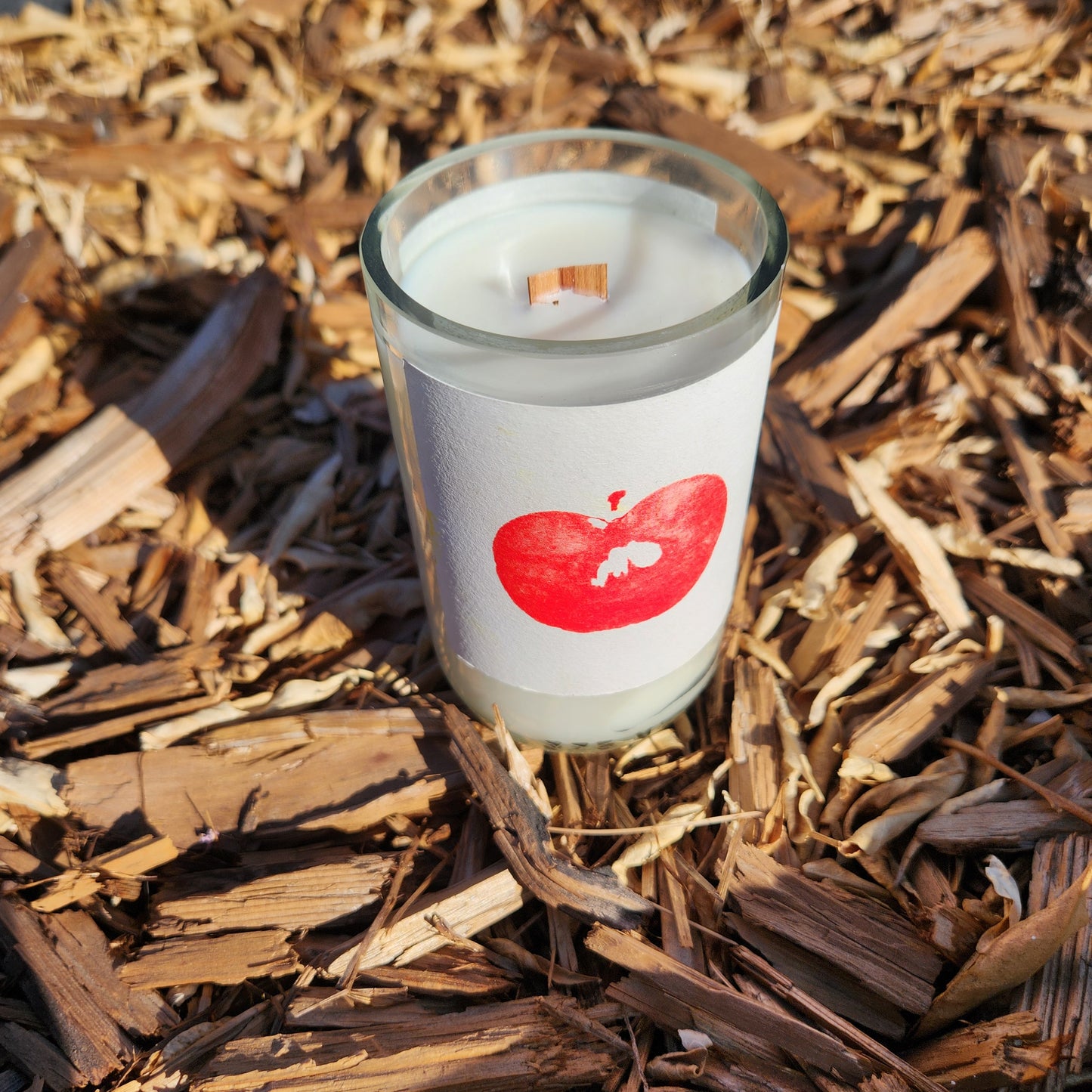 A hand-poured coconut-soy candle with a wooden wick in a recycled bottle, featuring a label with a minimalist red apple design. The candle is placed outdoors on a bed of wood chips, highlighting a natural and rustic aesthetic.