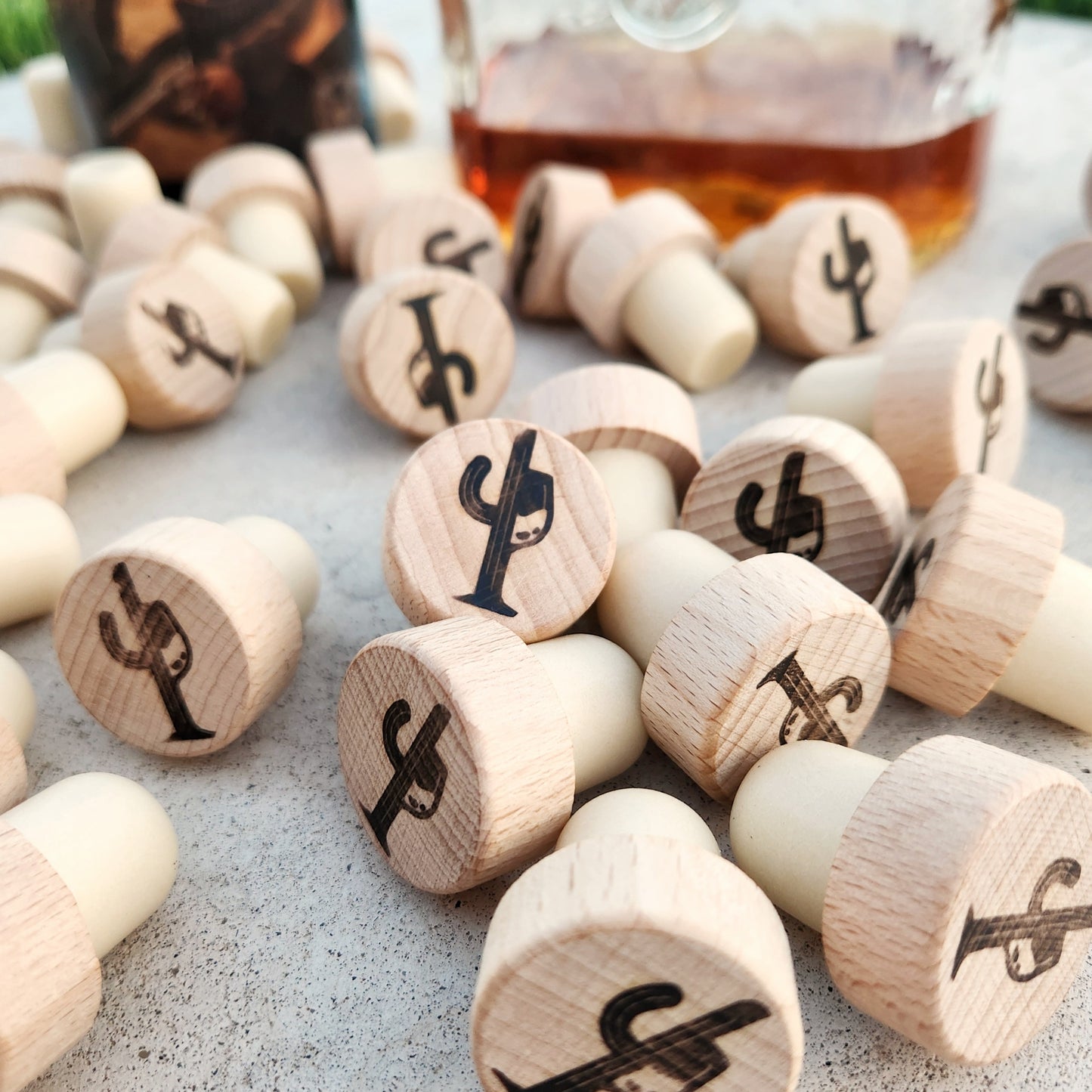 A collection of wooden bottle stoppers with natural cork bases spread across a concrete surface. Each stopper is engraved with a cactus and wine glass logo, showcasing a rustic, handcrafted design. A blurred glass bottle filled with amber liquid sits in the background, adding a touch of warmth and context.