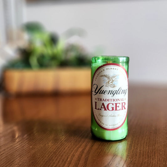 A recycled glass candle crafted from a green Yuengling Traditional Lager beer bottle, featuring the original label with red and gold accents and the iconic eagle logo. The candle sits on a polished wooden surface indoors, with a blurred background showcasing a wooden planter filled with green plants, creating a cozy and eco-friendly atmosphere.